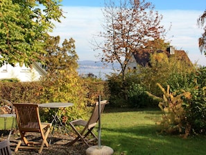 Ein Garten mit Tisch und Stühlen, einer Bank und Blick auf ein Gewässer sowie Gebäude in der Ferne.