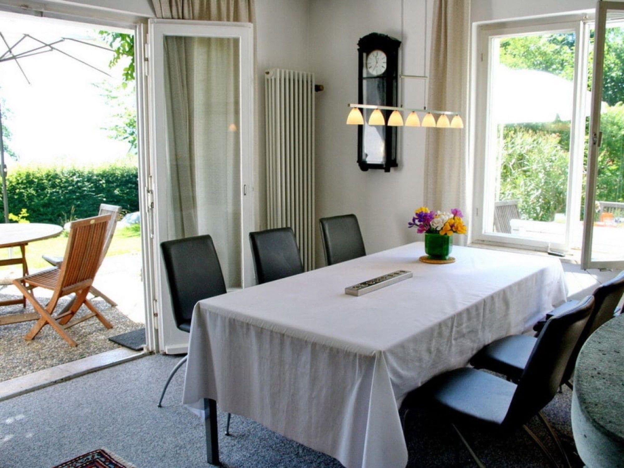 A dining area with a table set for four, chairs, a clock, and a window with a view of greenery.