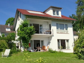 Ein zweistöckiges Haus mit rotem Ziegeldach, einem Balkon und einem kleinen Garten mit Stuhl und Tisch.