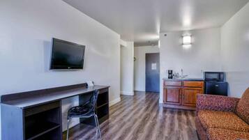 Quarto de hotel com escrivaninha, cadeira, televisão, cozinha compacta e sofá estampado.