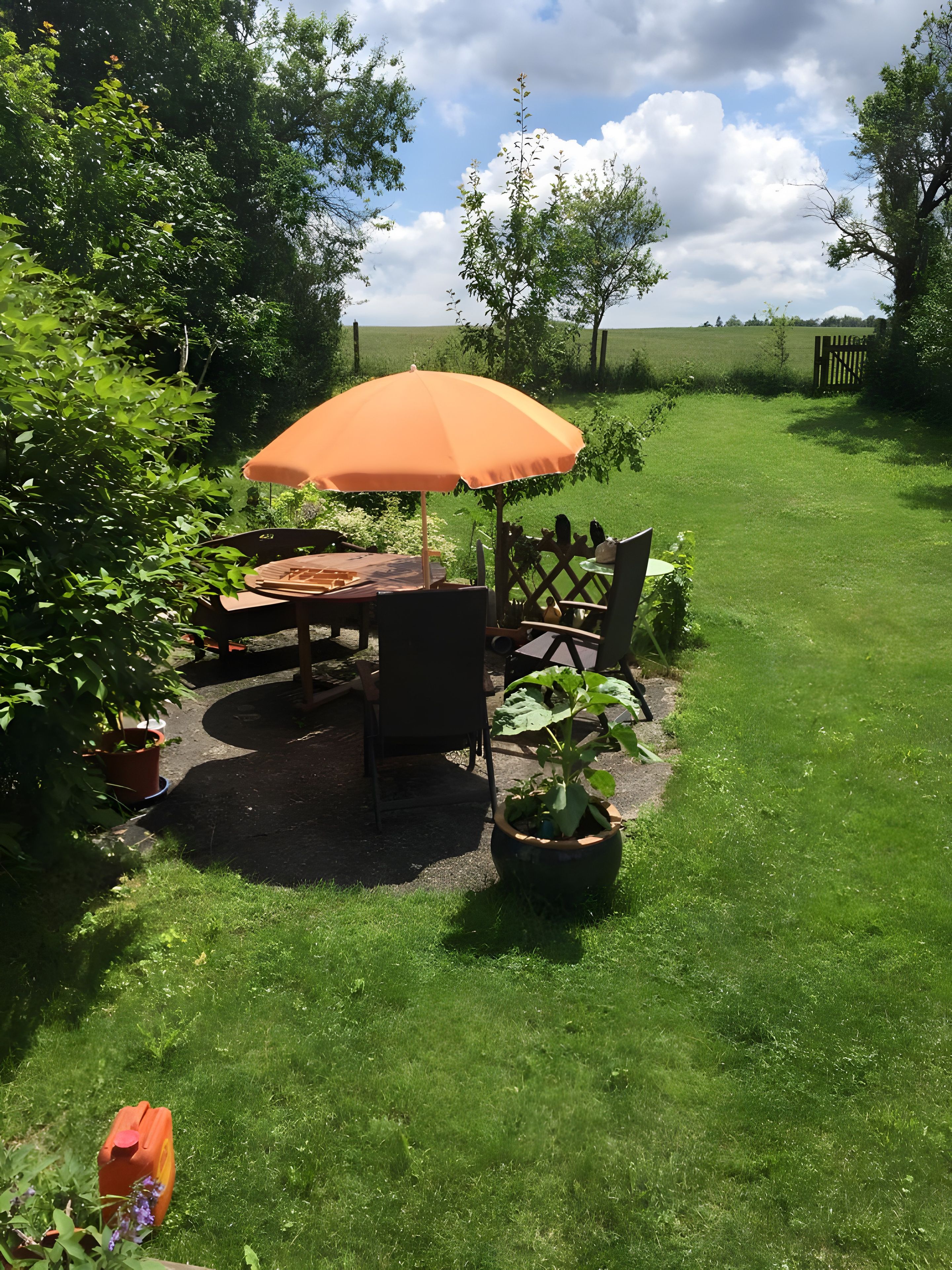 An outdoor patio area with a table and chairs under an umbrella, surrounded by greenery and a view of the countryside.