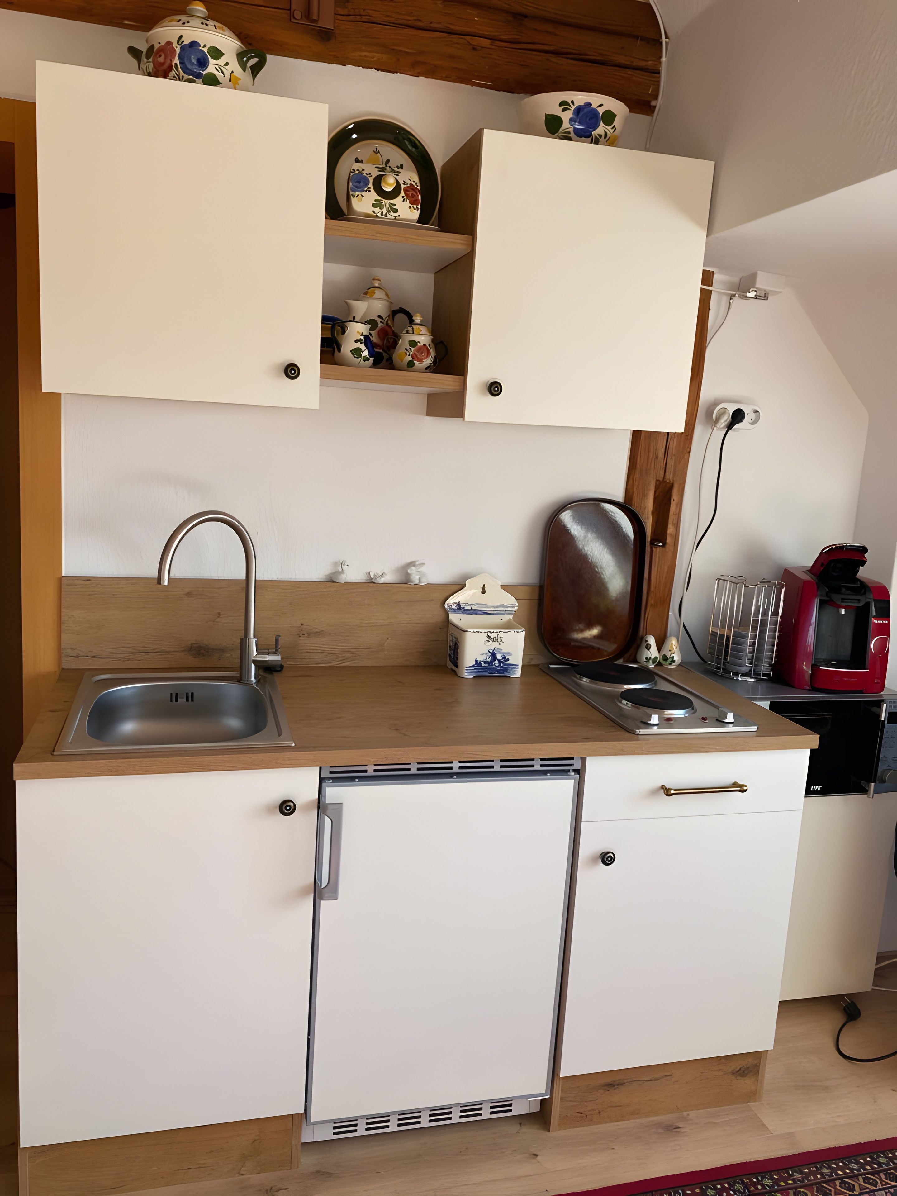 A compact kitchen with a sink, a compact refrigerator, and a microwave. There are cabinets above the counter, and a small shelf with decorative items.