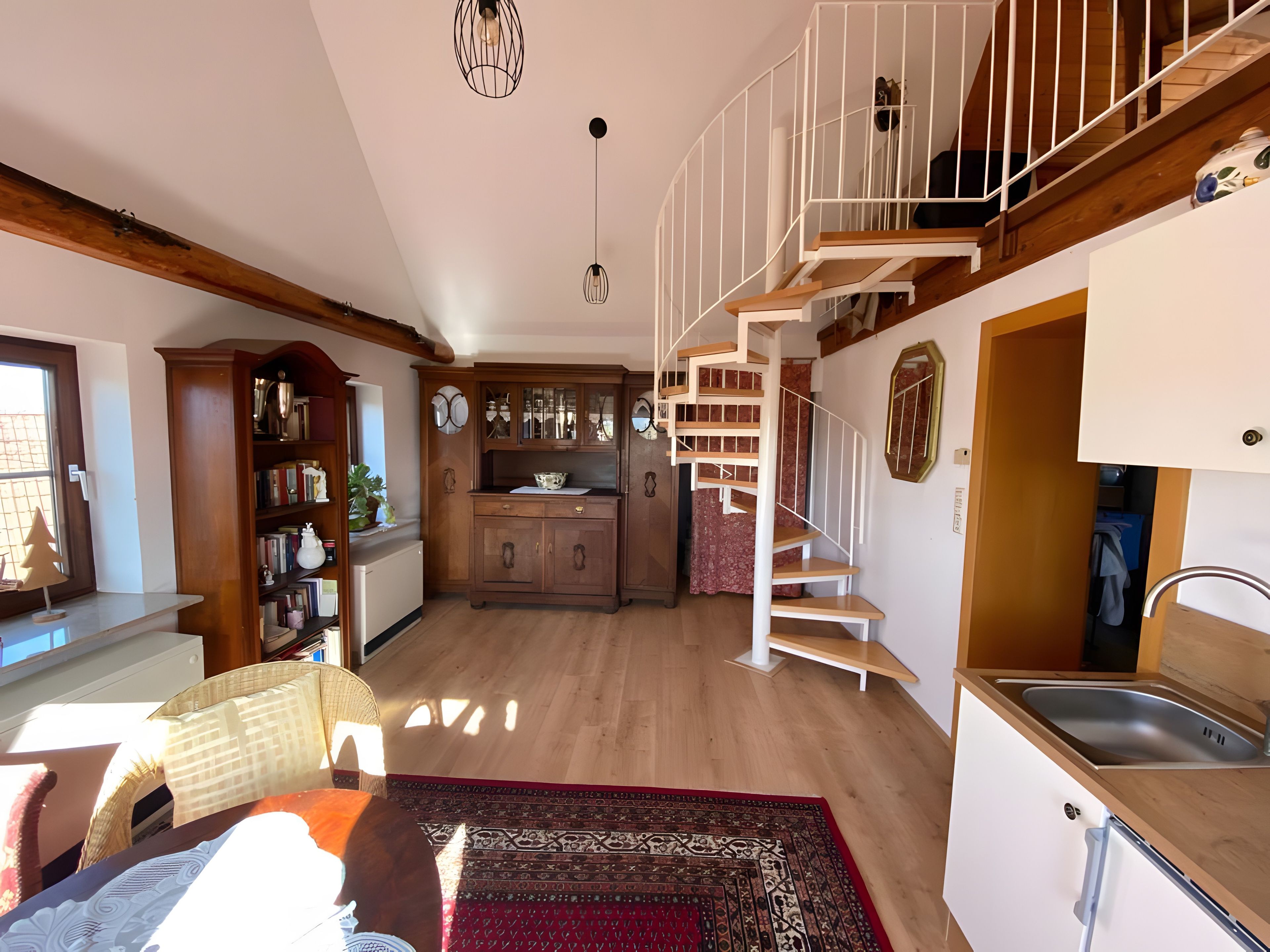 A spacious living area with a wooden staircase leading to a loft, a kitchen with wooden cabinets, and a dining area with a patterned rug.