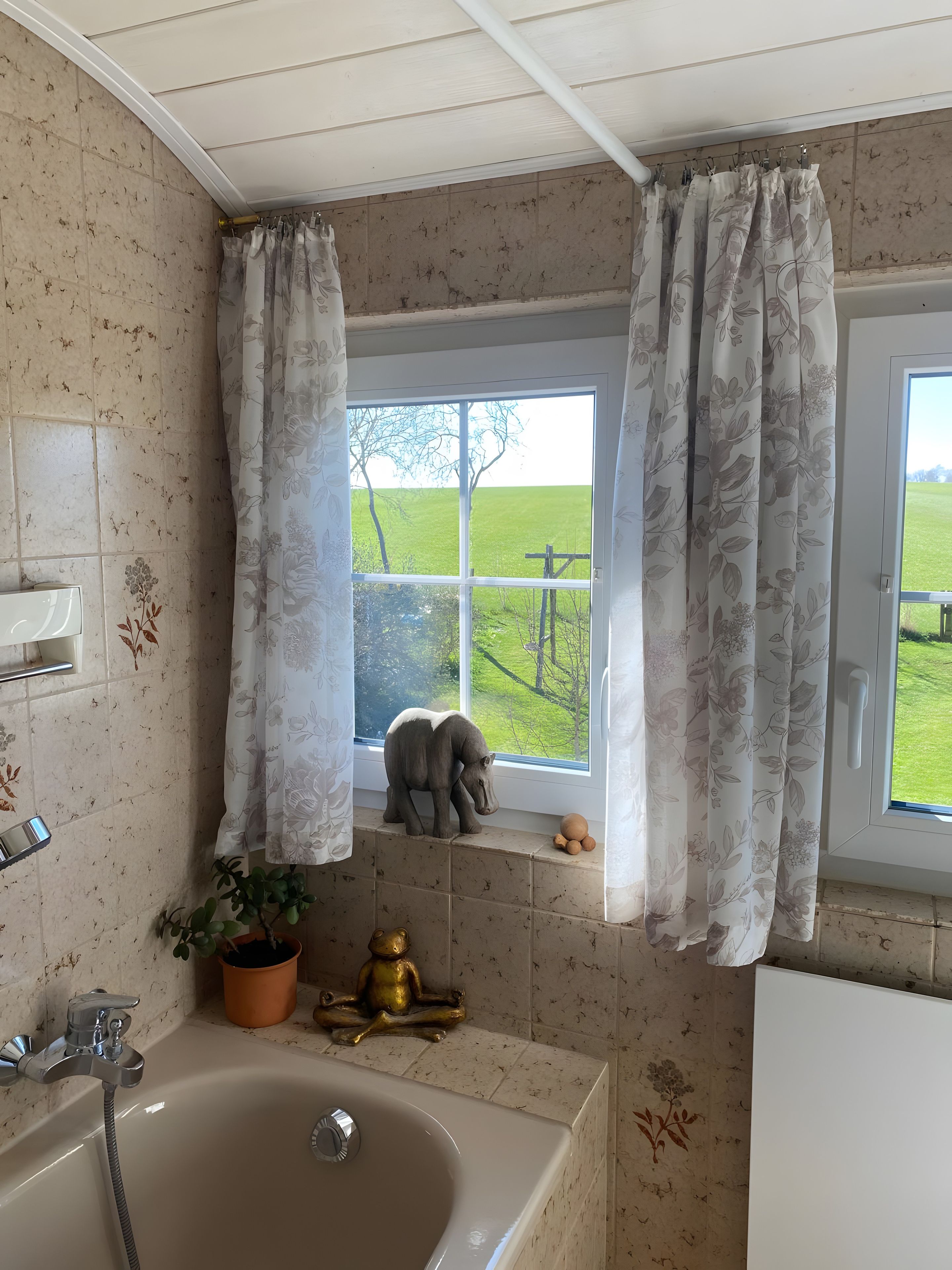 A bathroom with a bathtub, a window with curtains, a potted plant, and a small statue.