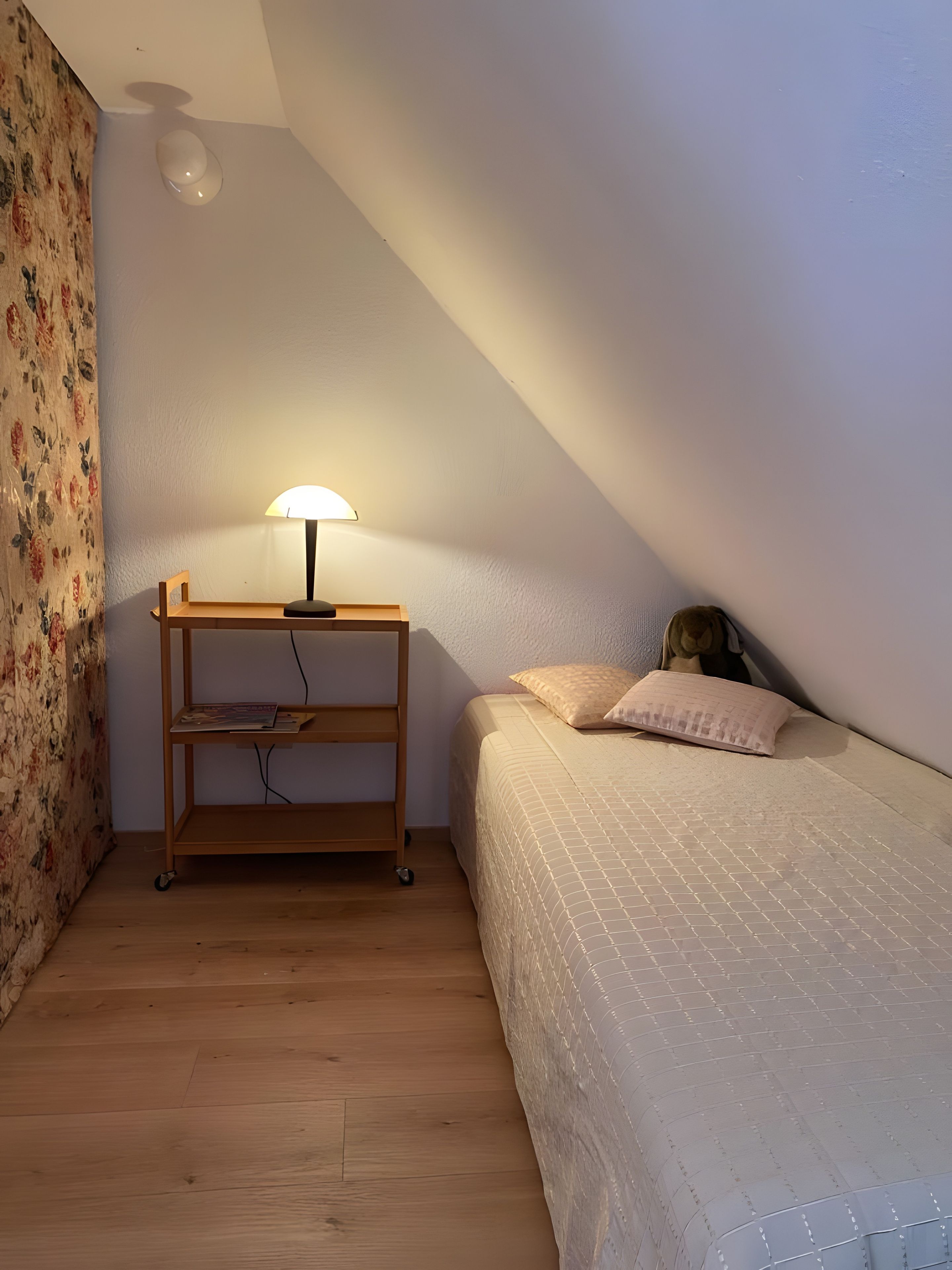 A bedroom with a wooden nightstand, a lamp, a bed with a quilt, and a floral curtain.
