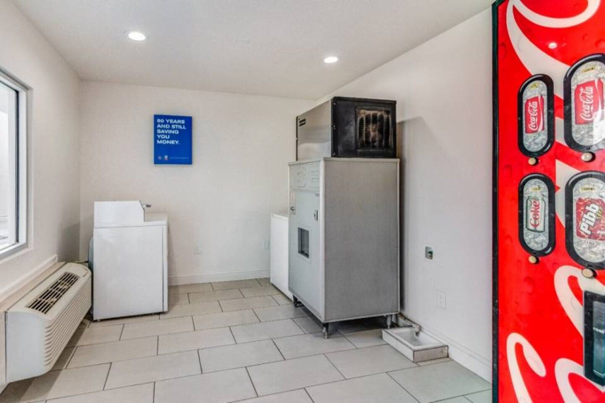 Eine kleine Küchenzeile mit einem Kühlschrank, einer Mikrowelle und einer Abluftheizung. An der Wand befindet sich ein blaues Schild und eine große Coca-Cola-Getränkeautomaten.