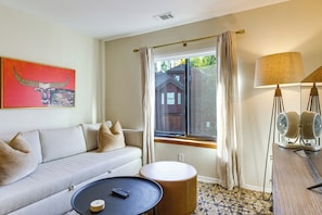 Ein Zimmer mit einer Couch, einem runden Hocker, einem kleinen Tisch mit Fernbedienung, einer Lampe und einem Fenster mit Vorhängen.