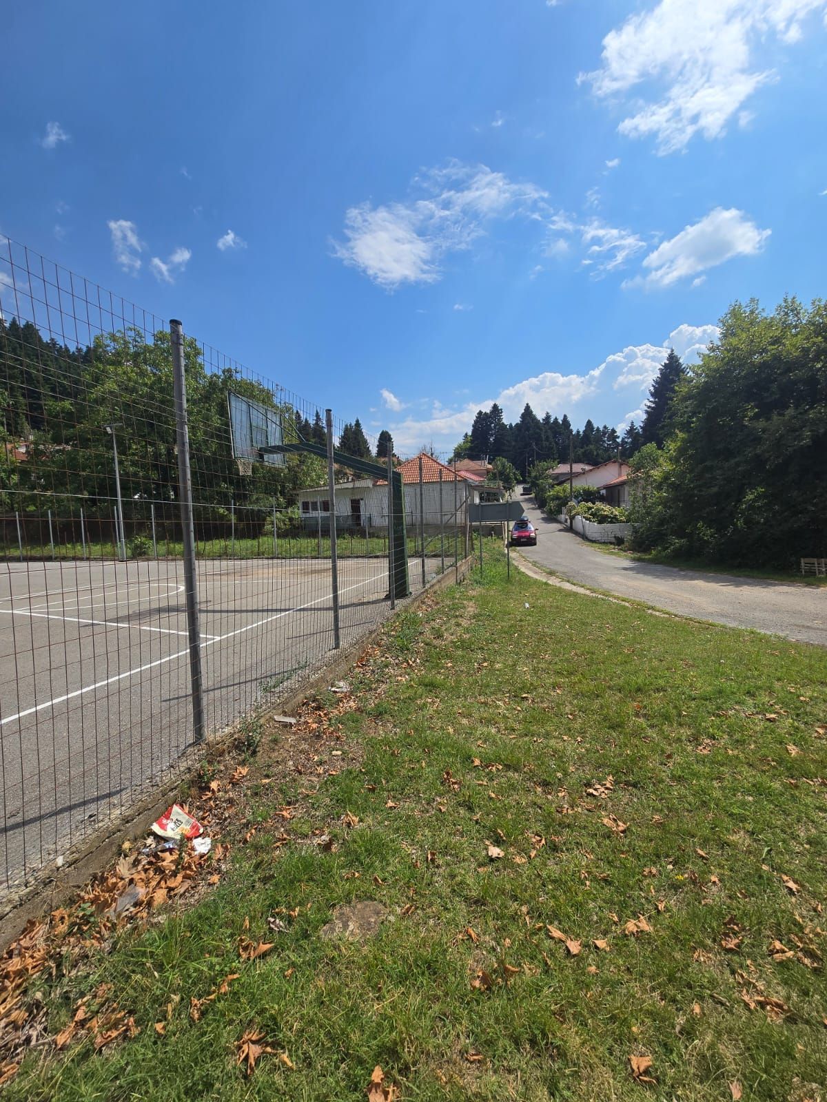 Een omheind sportterrein met een weg naar huizen, omringd door bomen en een heldere lucht.