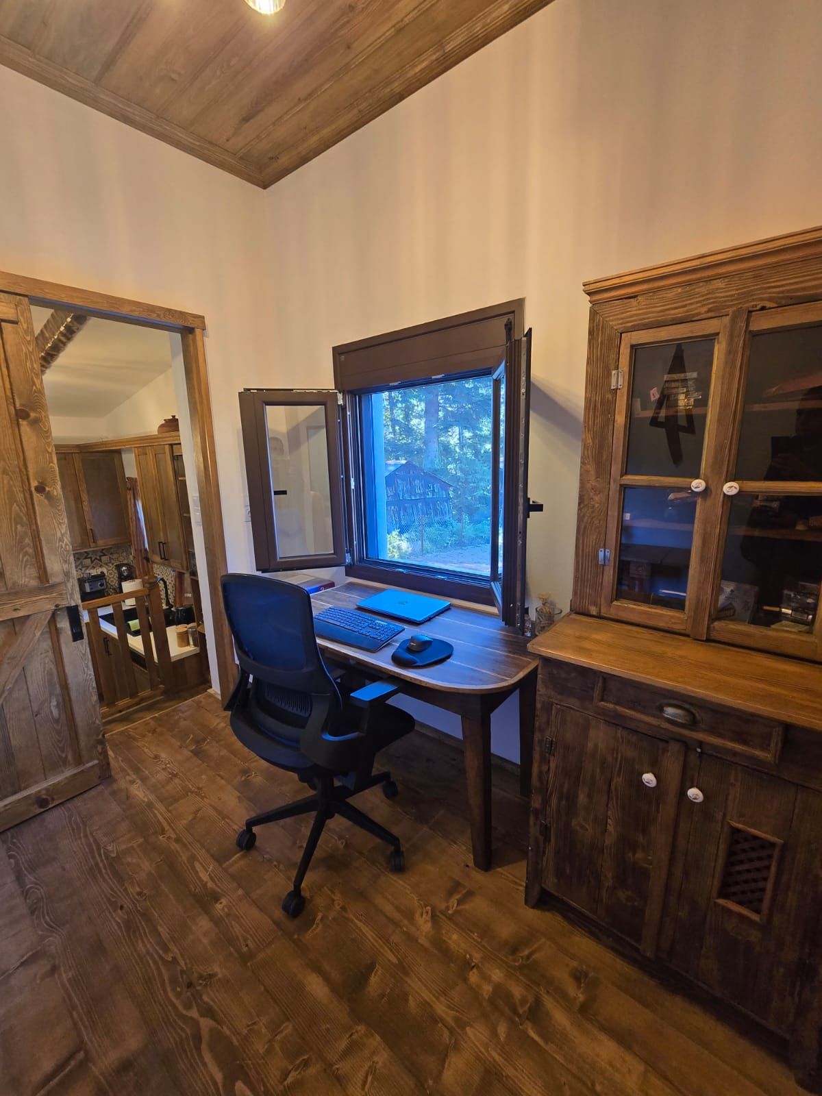 A wooden-paneled room with a desk, chair, and a window overlooking a forest.