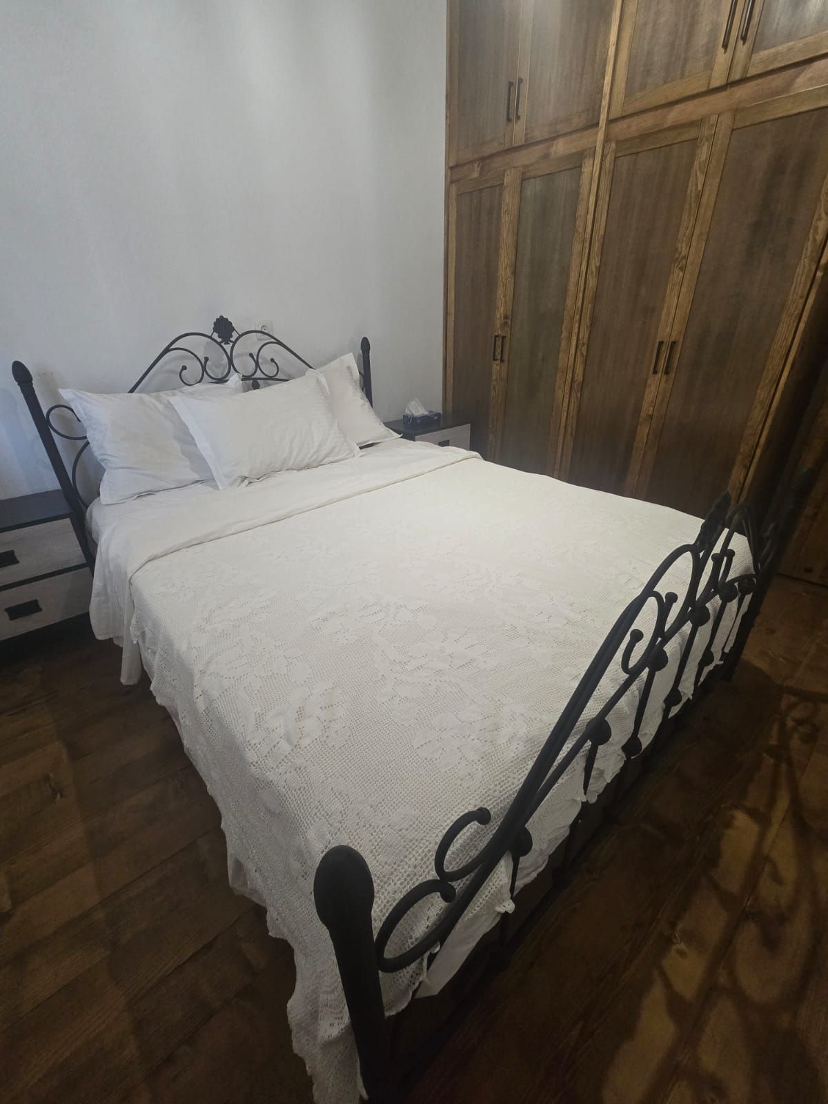 Un letto singolo con struttura in metallo e biancheria da letto bianca, un armadio in legno e un comodino con una lampada.