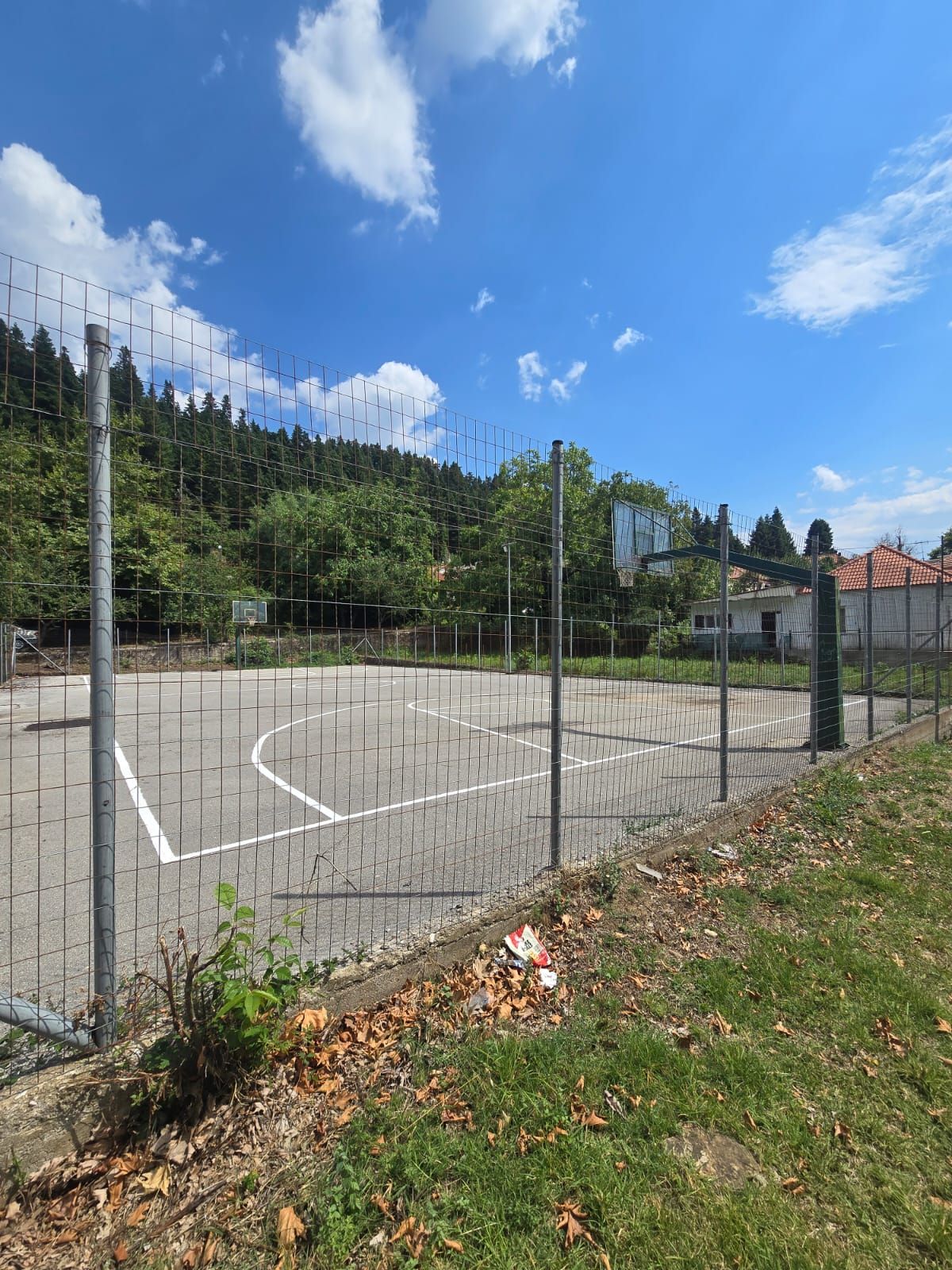 An outdoor basketball court enclosed by a metal fence, surrounded by trees and a clear sky.
