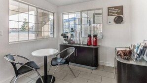 Ein kleiner, moderner Raum mit einem weißen Tisch und zwei Stühlen, einem Fenster mit Blick nach draußen und einer Theke mit einer Kaffeemaschine und einem Schild.