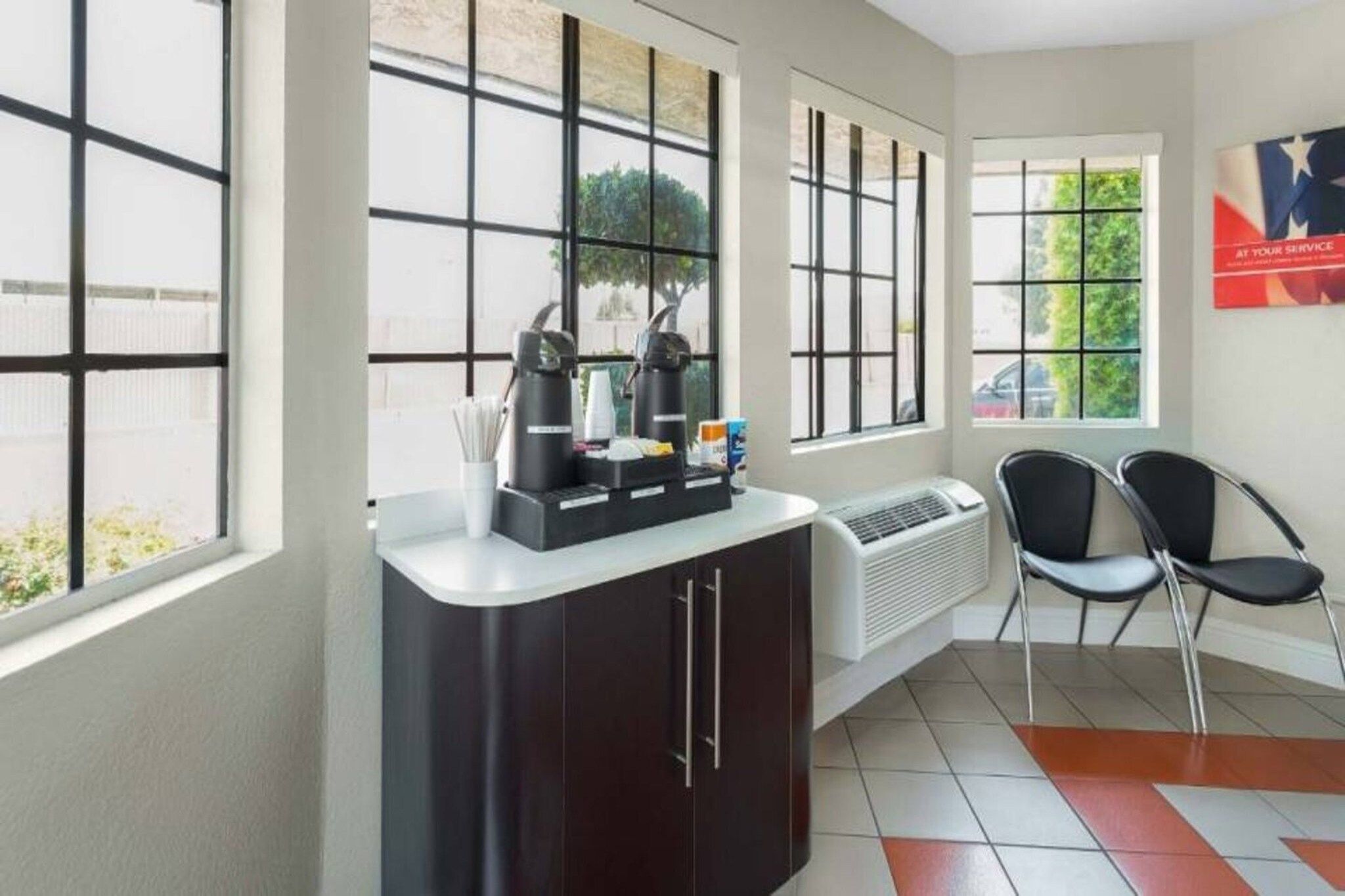 A hotel room with a kitchenette, a seating area with chairs, large windows, and a poster on the wall.