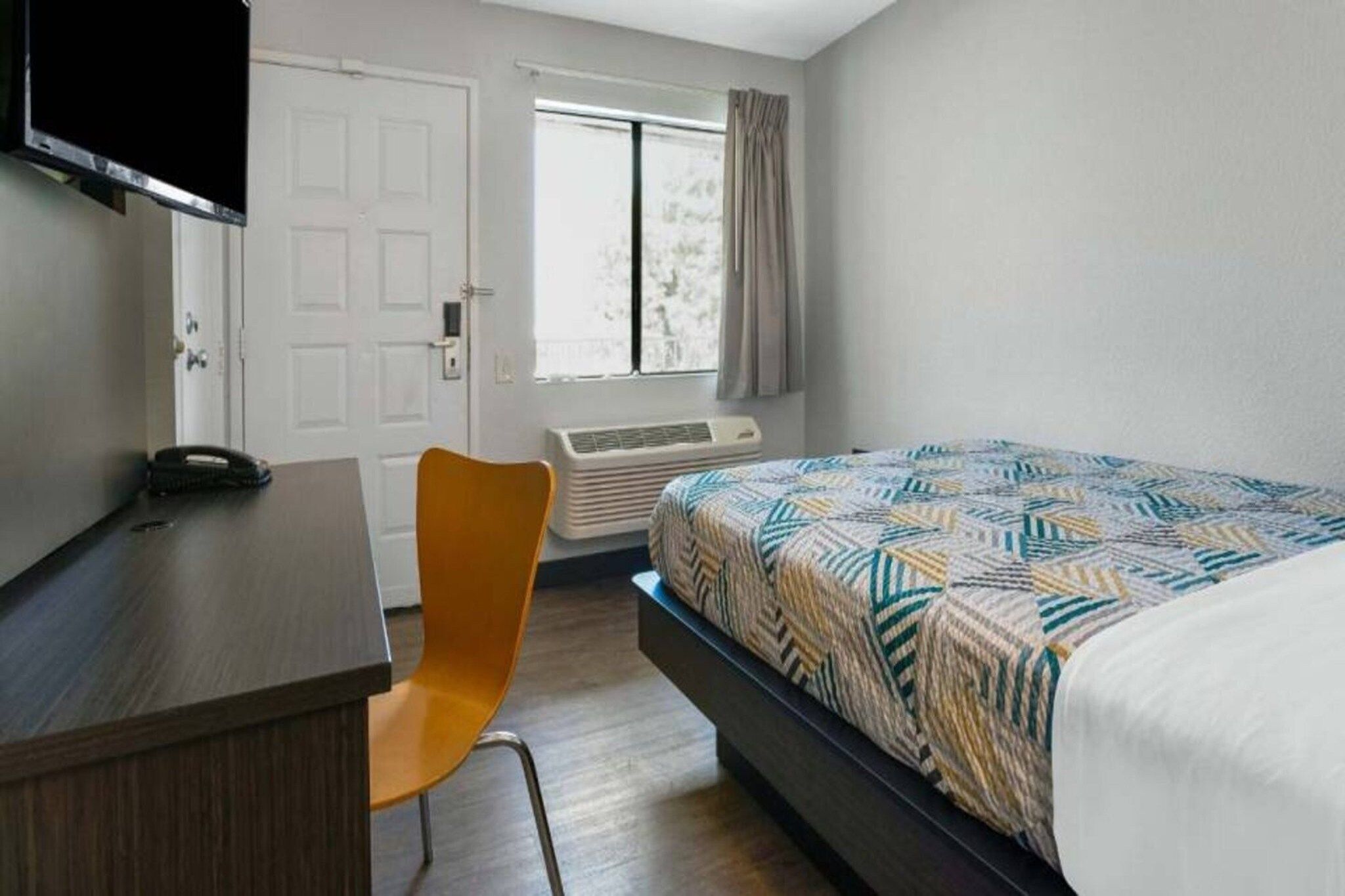 Une chambre à coucher comprenant un lit, un bureau, une chaise, un téléviseur, une fenêtre avec des rideaux et un appareil de climatisation.