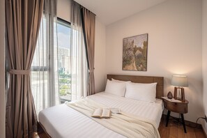Habitació d'hotel amb un llit gran, una tauleta de nit amb una làmpada, un quadre a la paret i vista a la ciutat a través de cortines primes.