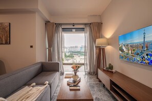 A modern living room with a grey sofa, a coffee table with books and a vase, a lamp, and a large window with curtains. - Spacious 3BR Family Stay in Dist 1 _ Pool, Gym & Balcony (Thành phố Hồ Chí Minh)
