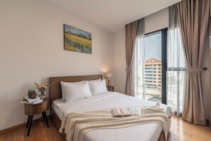 A hotel room with a bed, a nightstand with books and a vase, a painting on the wall, and a view through the window. - Spacious 3BR Family Stay in Dist 1 _ Pool, Gym & Balcony (Thành phố Hồ Chí Minh)