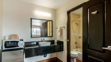 A bathroom with a sink, mirror, toilet, shower, and a microwave on a counter.