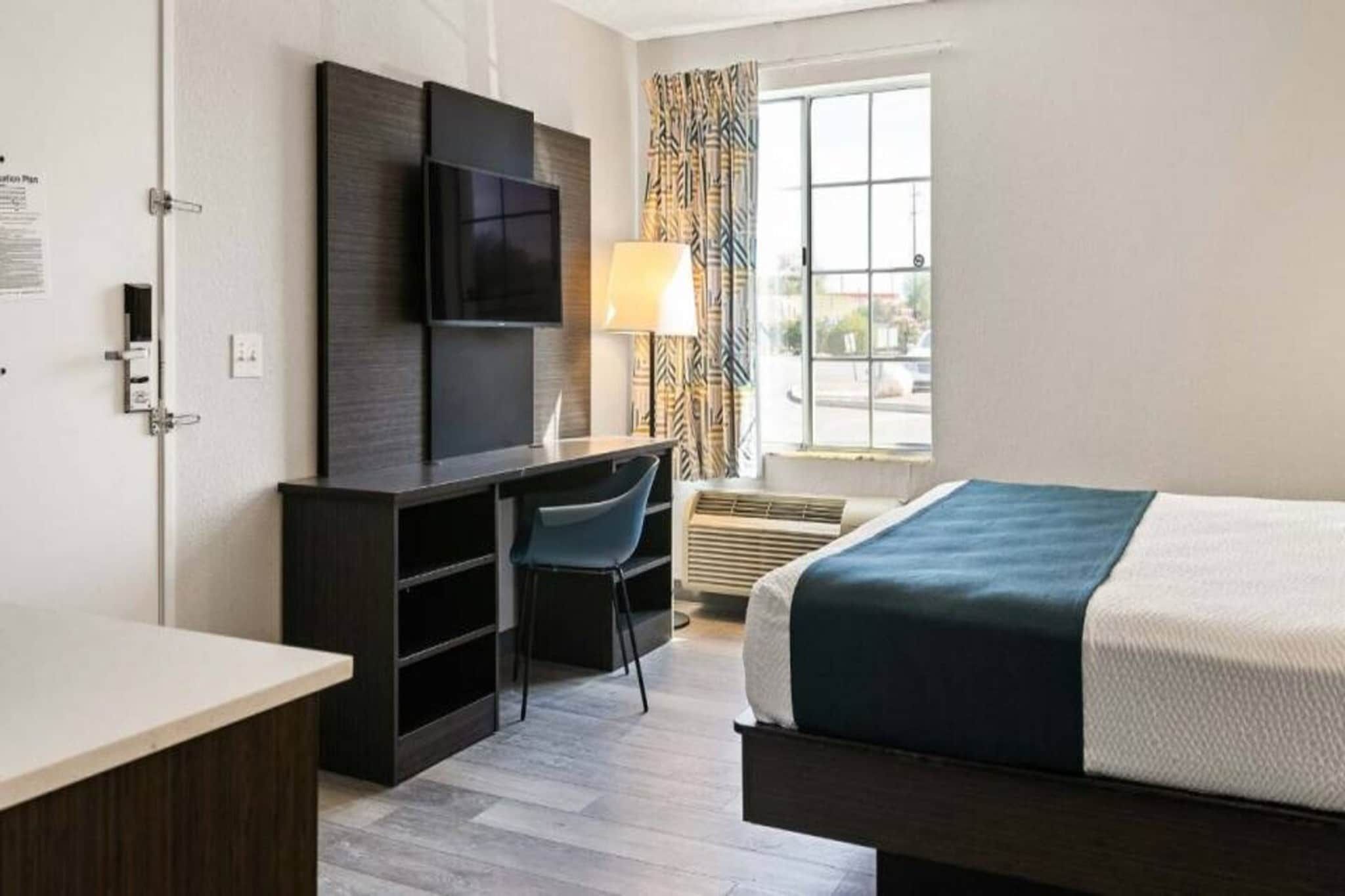 Habitación de hotel con cama, escritorio, silla, televisor y ventana con cortinas.