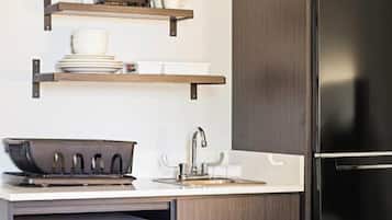 A kitchen with a stainless steel refrigerator, a sink, and a stove. There are wooden shelves above the counter holding dishes and cups.