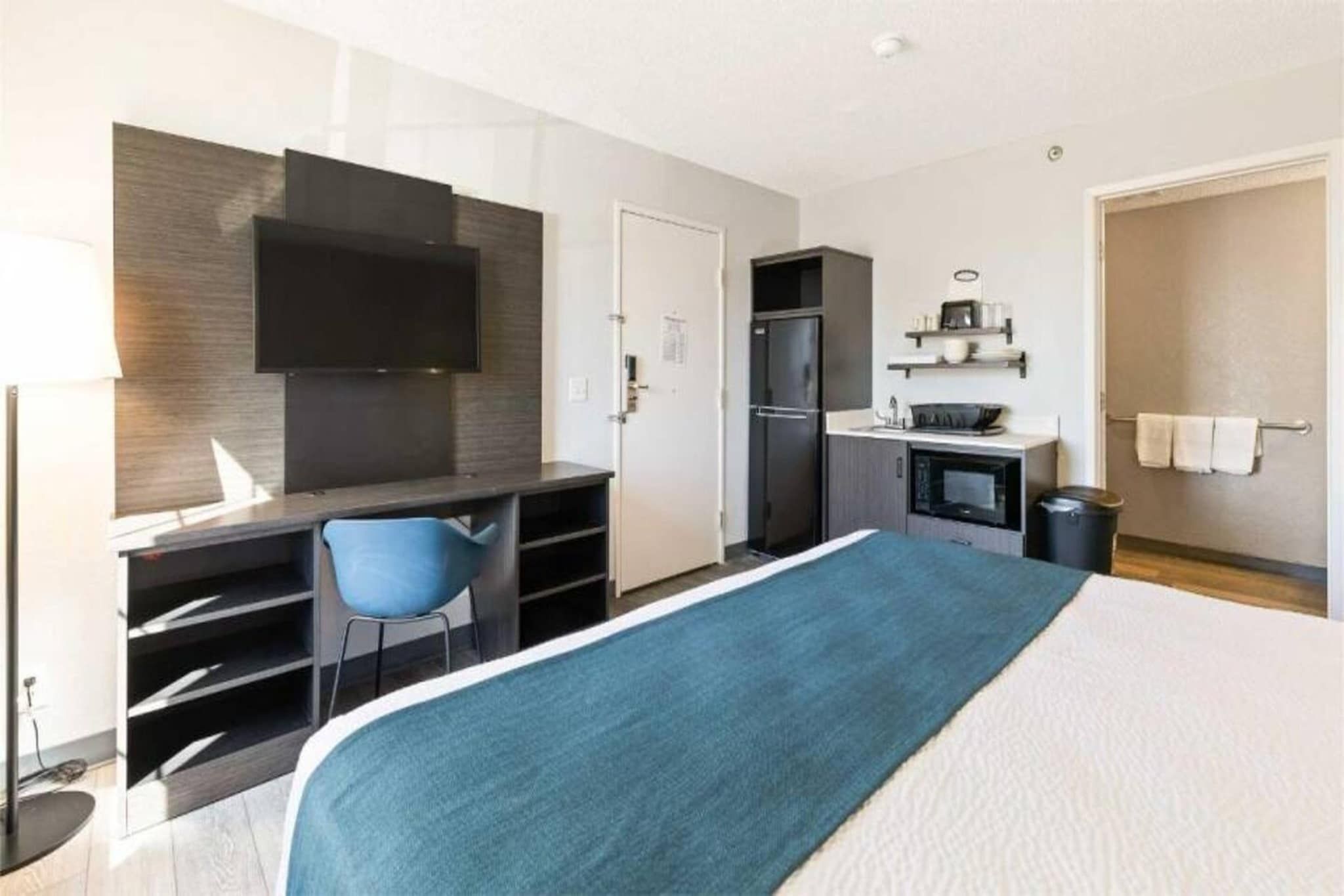 A hotel room with a bed, a desk, a chair, a TV, a kitchenette, and a bathroom.