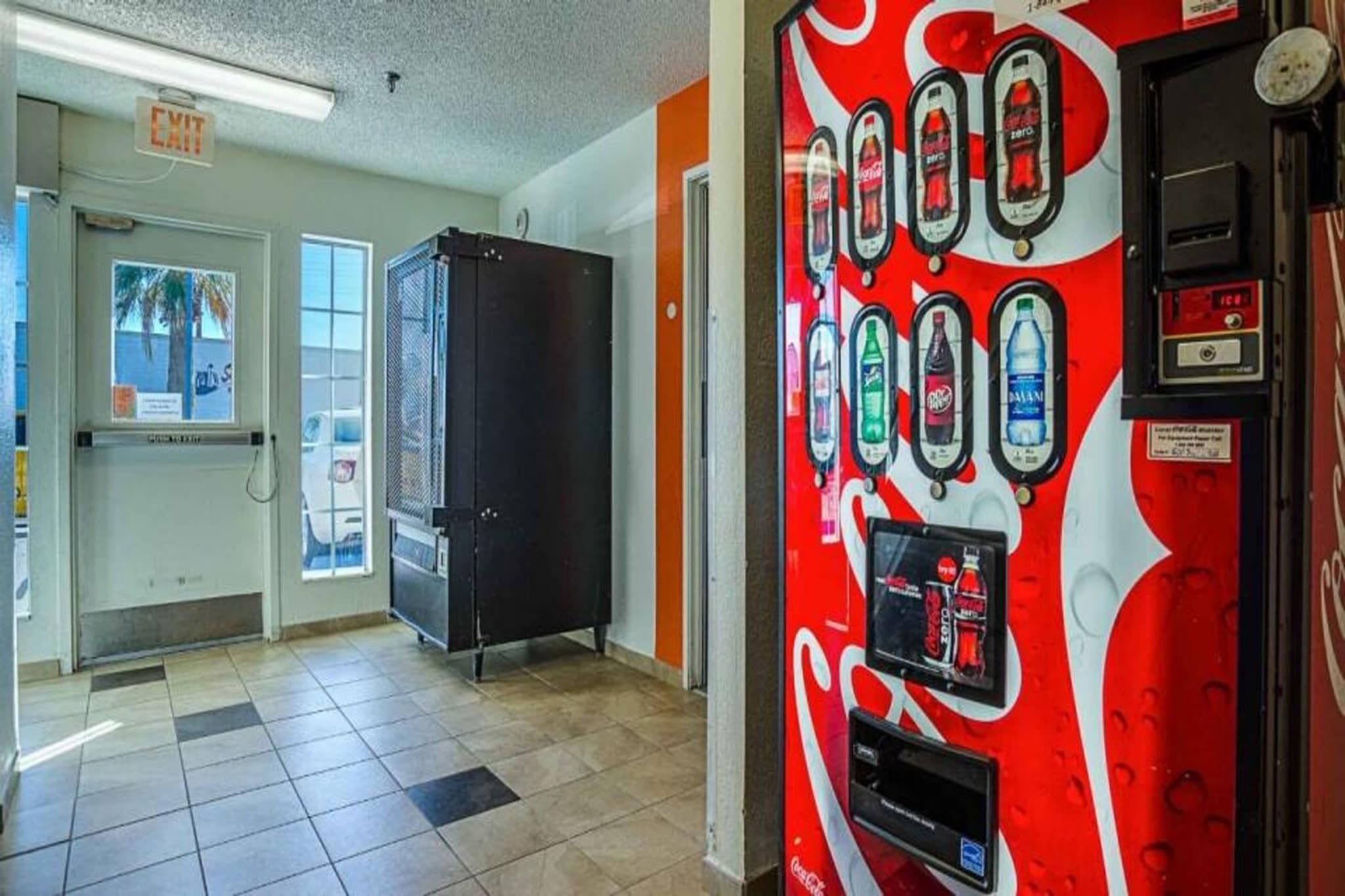 A vending machine with various drink options, a door with an exit sign, and a window with a view of palm trees.