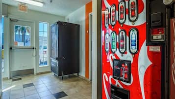 A vending machine with various drink options, a door with an exit sign, and a window with a view of palm trees.