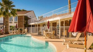 Ein Schwimmbecken mit Liegestühlen und Sonnenschirmen, ein Gebäude mit Balkon und eine Palmenbaum.