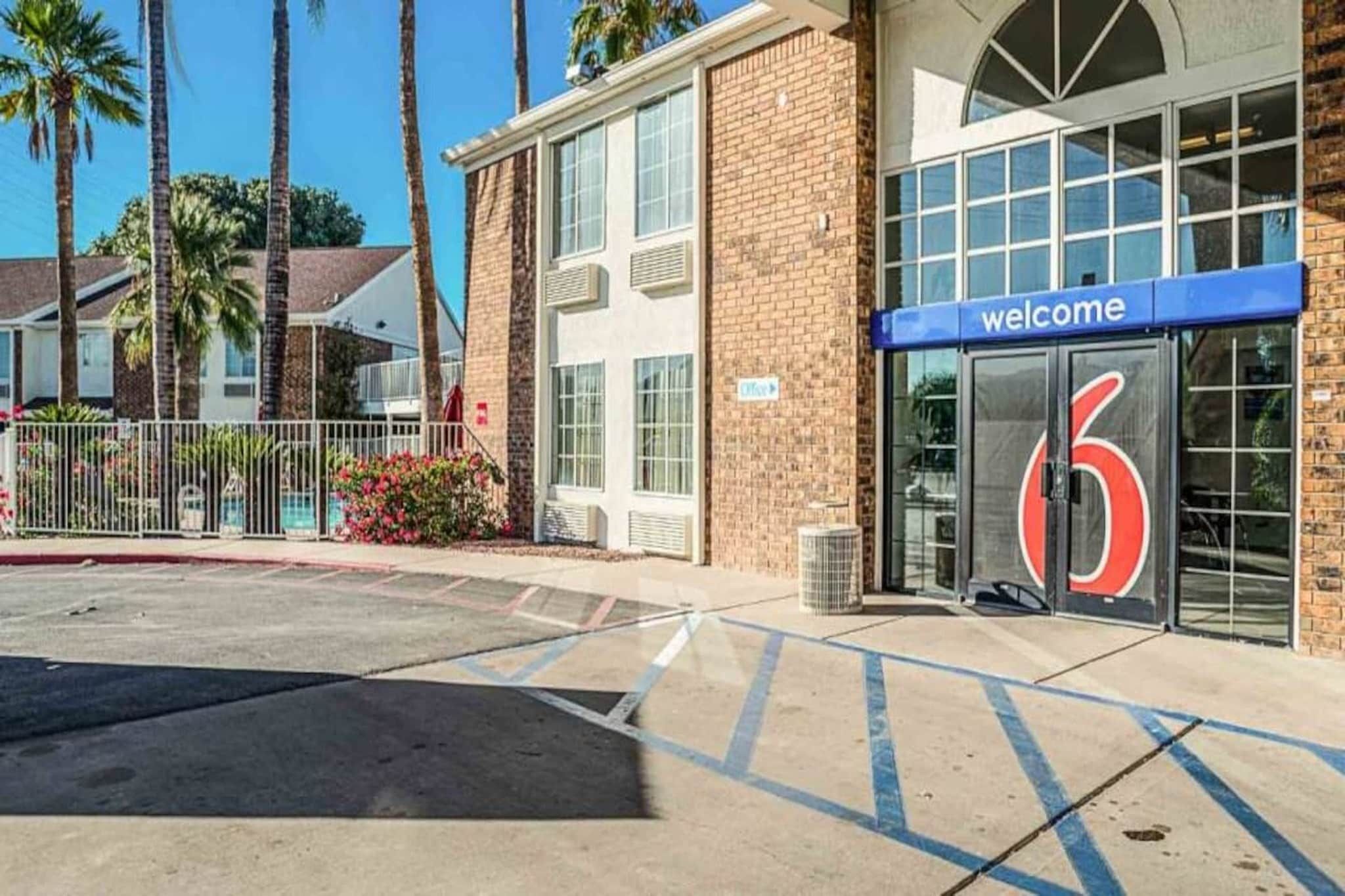 Ein Hotel Eingang mit einer '6' Beschilderung, einem 'Willkommen'-Banner und einem Parkplatz mit markierten Stellplätzen.