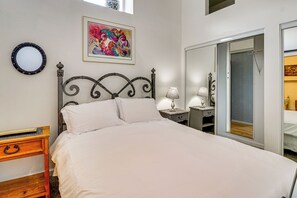 Un dormitorio con una cama, una mesita de noche con lámpara, un aparador, un espejo y un cuadro de colores en la pared.