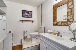Un baño con inodoro, lavamanos, espejo y repisa con una imagen sobre el inodoro.