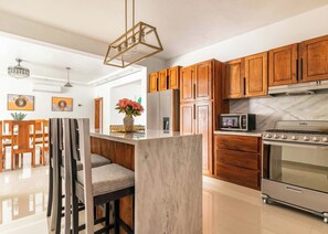 A modern kitchen with wooden cabinets, a stainless steel refrigerator, a built-in oven, and a bar area with stools. - 10BR Luxury Villa  Private Pool (Sosúa)