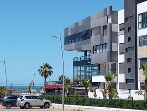 A modern multi-story building with a glass facade, located near a beach with palm trees and a parked car. - Relaxing Stay with Pool  Garden Near Mohammedia Beach (El Mansouria)