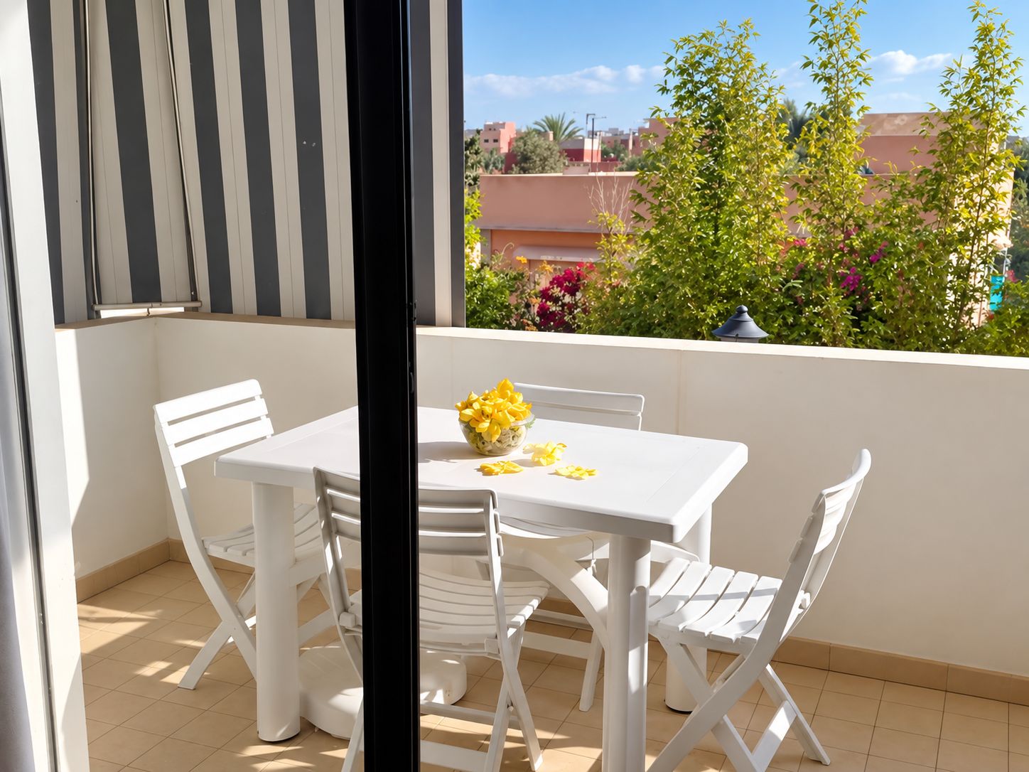 Un balcon avec une table et des chaises blanches, un vase rempli de fleurs jaunes et une vue sur des bâtiments et de la verdure.
