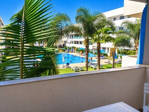 Un área de piscina con sillones de descanso y palmeras, rodeada por un edificio de varios pisos.