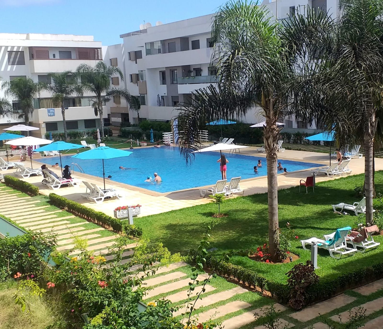 A swimming pool with lounge chairs and umbrellas, surrounded by greenery and buildings.