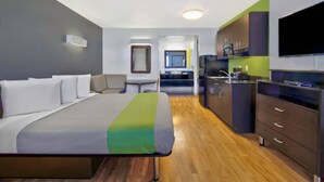 A modern hotel room with a bed, a small kitchenette, a flat-screen TV, and a bathroom with a mirror and sink. - MS6 Victorville|1 King Bed|NS (VICTORVILLE)