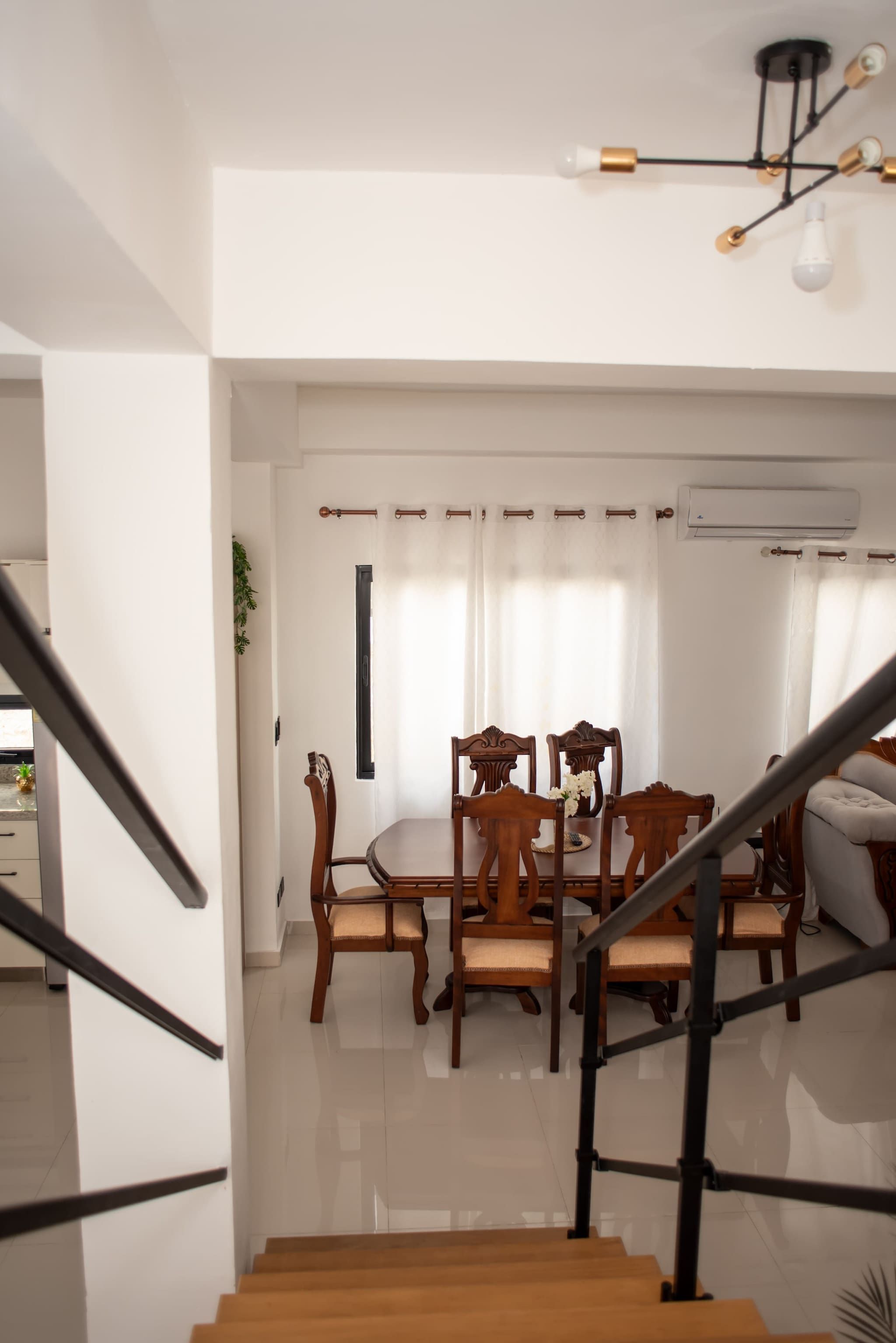 A staircase leading to a dining area with wooden chairs and a table, an air conditioning unit on the wall, and a ceiling light fixture.
