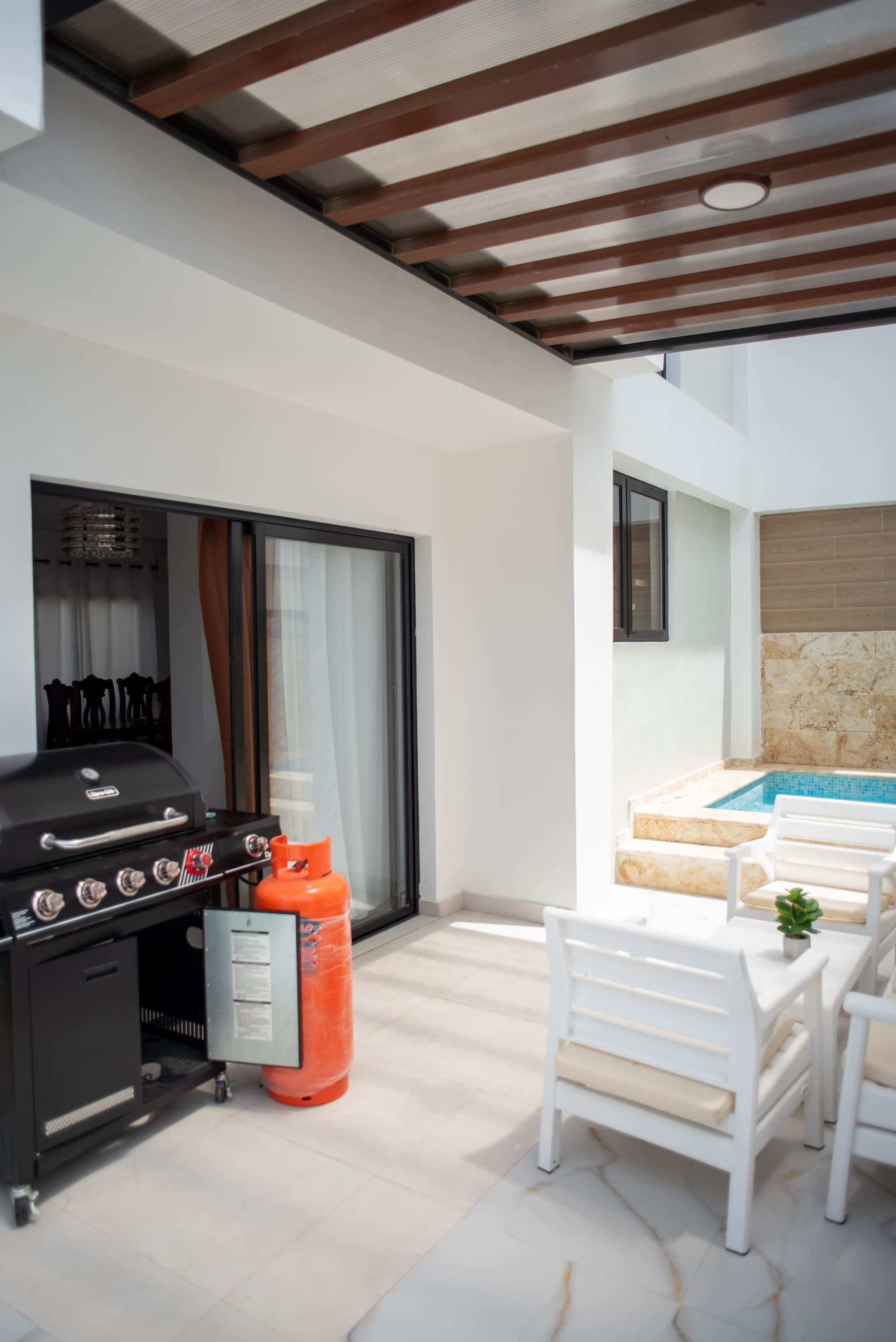 A modern outdoor patio with a barbecue grill, a gas cylinder, white outdoor furniture, and a swimming pool.