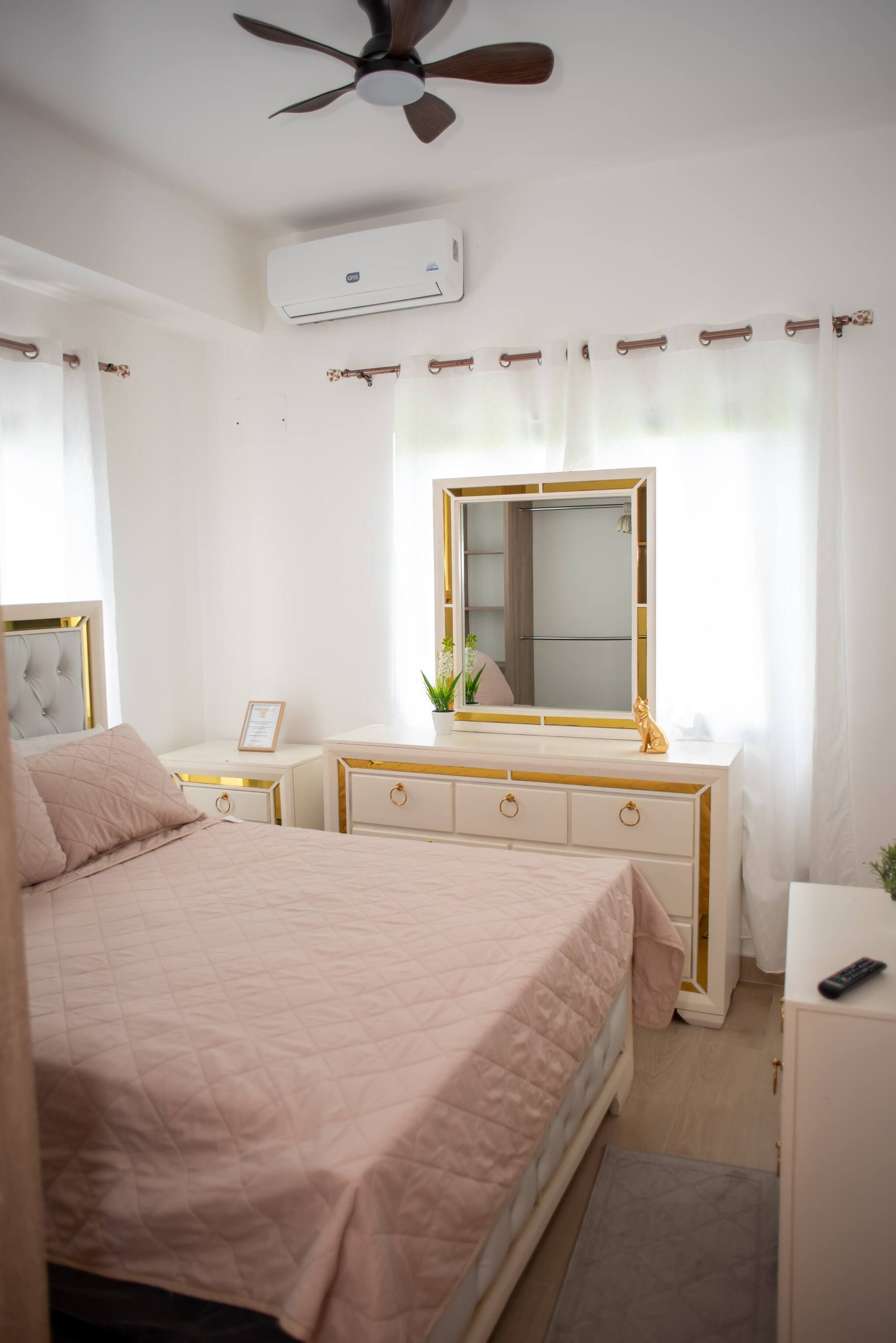 A bedroom with a bed, a vanity with a mirror, a ceiling fan, an air conditioning unit, and a window with curtains.
