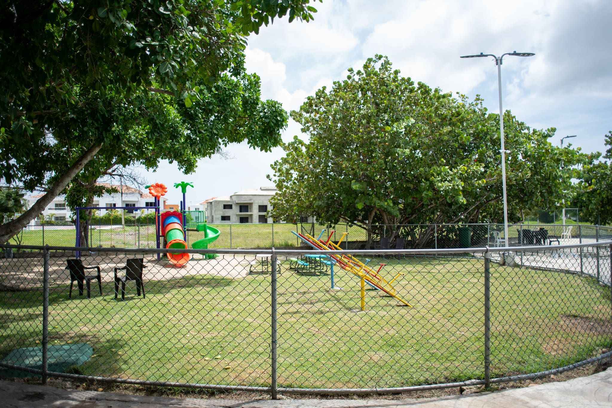 Un'area giochi recintata con diverse attrezzature, tra cui uno scivolo e altalene, circondata da alberi e un edificio sullo sfondo.