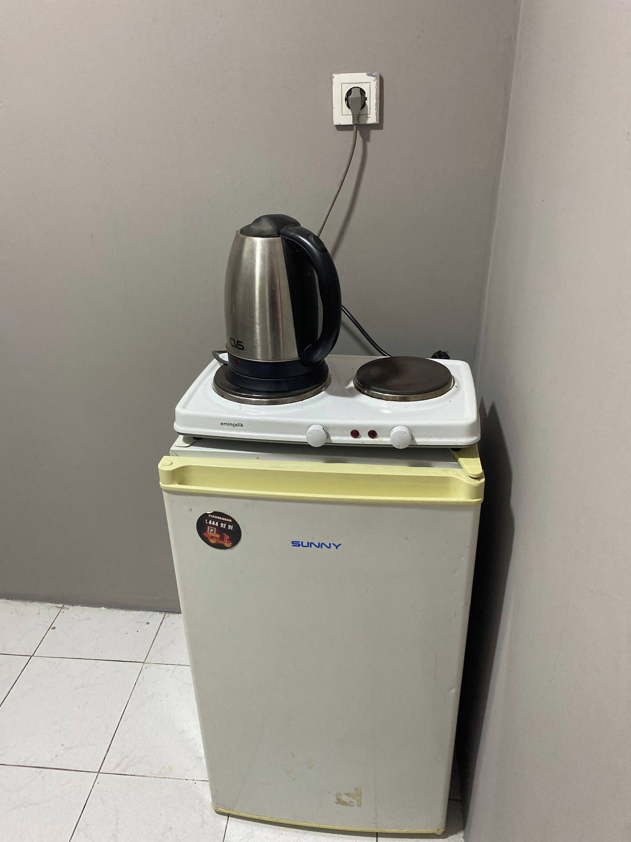 A compact refrigerator with a kettle on top, a power outlet on the wall, and a sticker with a phone number.