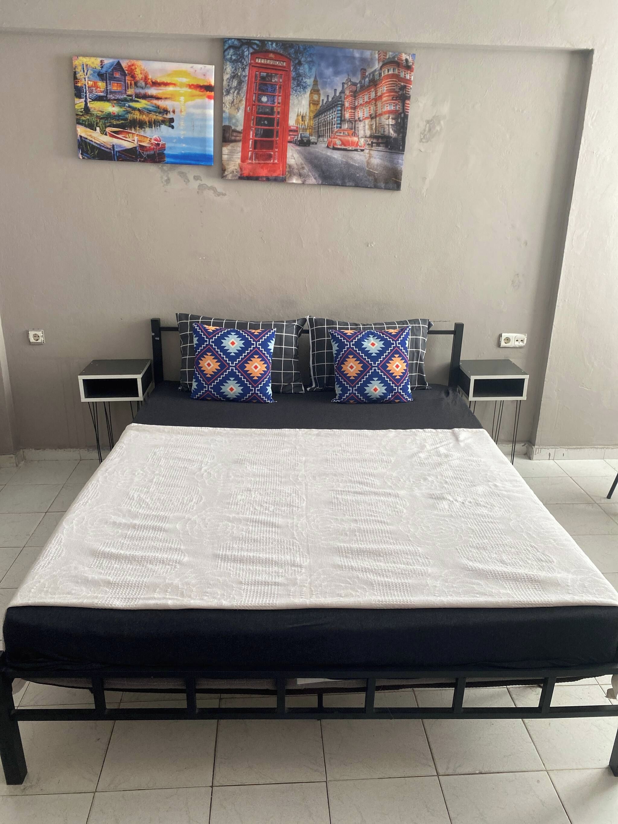 Ein Bett mit einem gemusterten Überwurf, zwei Kissen mit geometrischen Mustern und zwei Nachttische mit Lautsprechern.