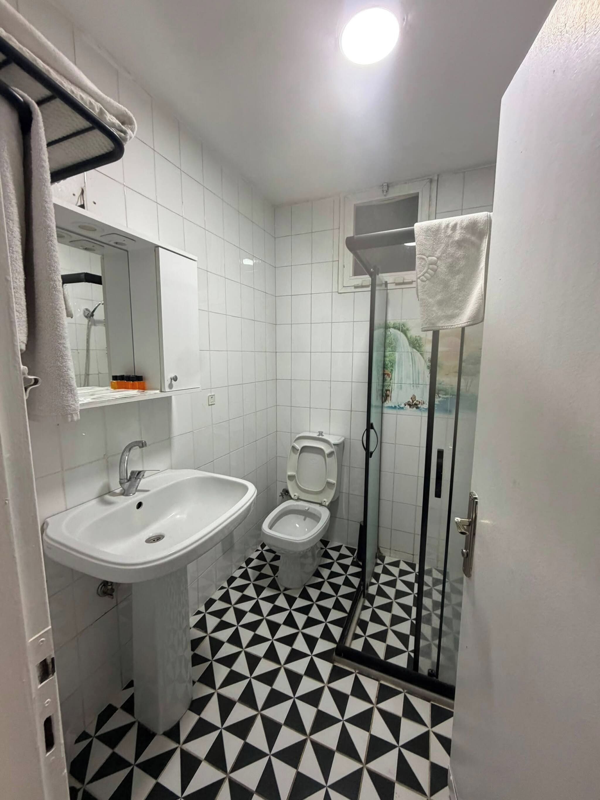 Ein Badezimmer mit weißem Waschbecken, WC und Dusche. Der Boden weist ein schwarz-weißes geometrisches Muster auf.