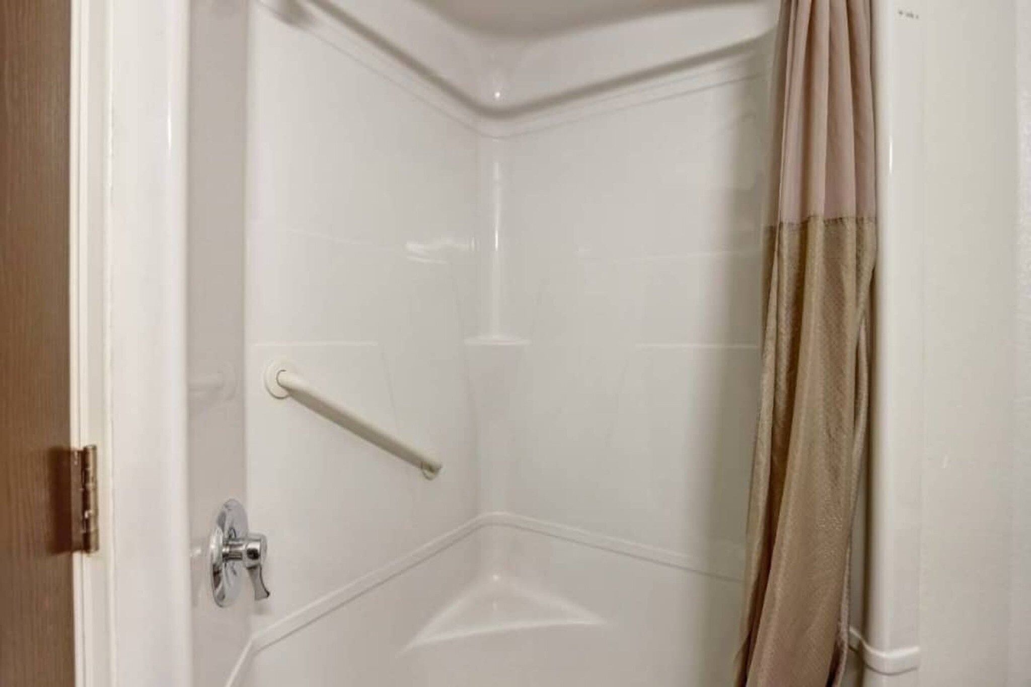 A corner shower with a white tiled wall, a metal grab bar, and a beige shower curtain.