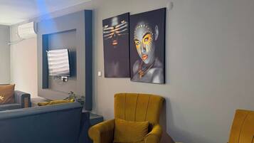 Una sala moderna con una lámpara de techo azul, dos sillones color mostaza, un reposapiés gris y dos cuadros abstractos en la pared.