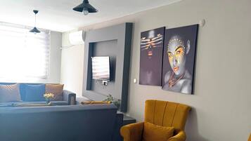 A modern living room with a sofa, ottoman, and two armchairs, a flat-screen TV mounted on the wall, and artwork above the seating area.