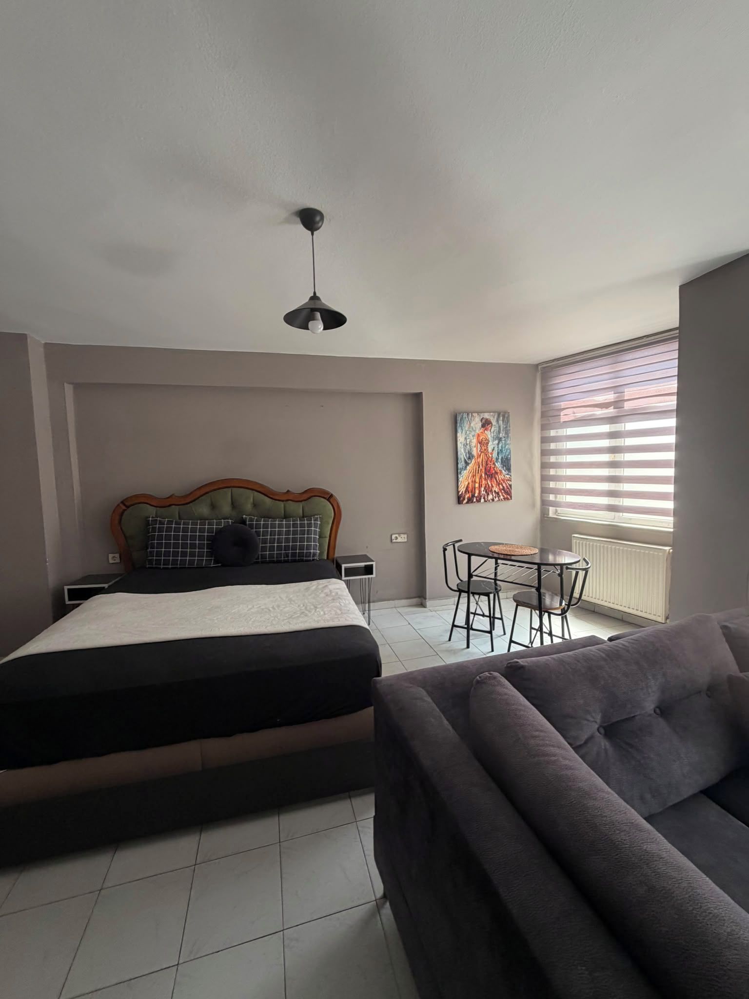 Ein Schlafzimmer mit einem Bett, einem Sofa, einem Esstisch mit Stühlen, einer Deckenleuchte und einem Fenster mit Jalousien.