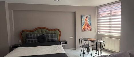 Ein Schlafzimmer mit einem Bett, einem Sofa, einem Esstisch mit Stühlen, einer Deckenleuchte und einem Fenster mit Jalousien.