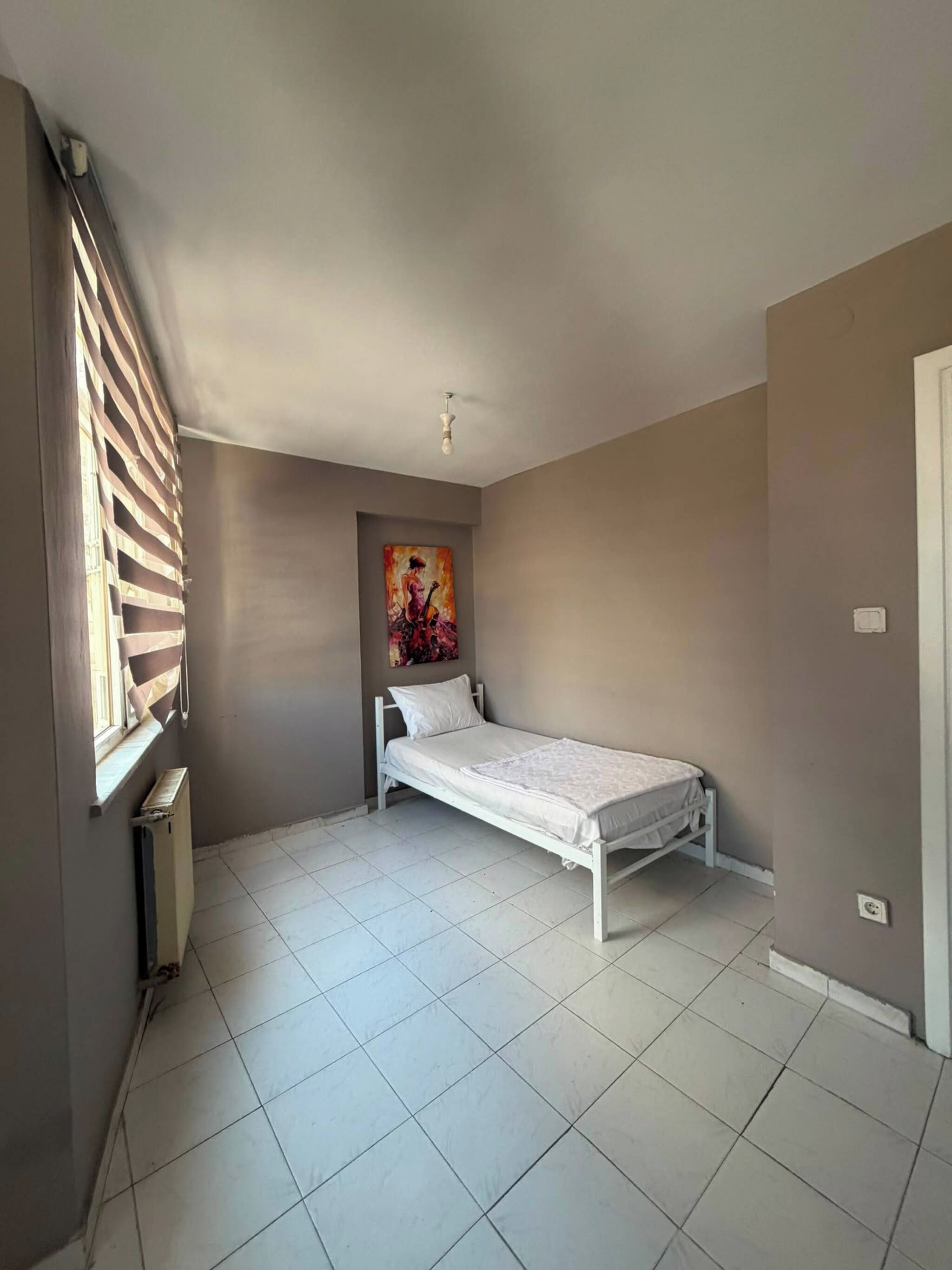 A small, single-bed room with a radiator, a window with blinds, and a painting on the wall.